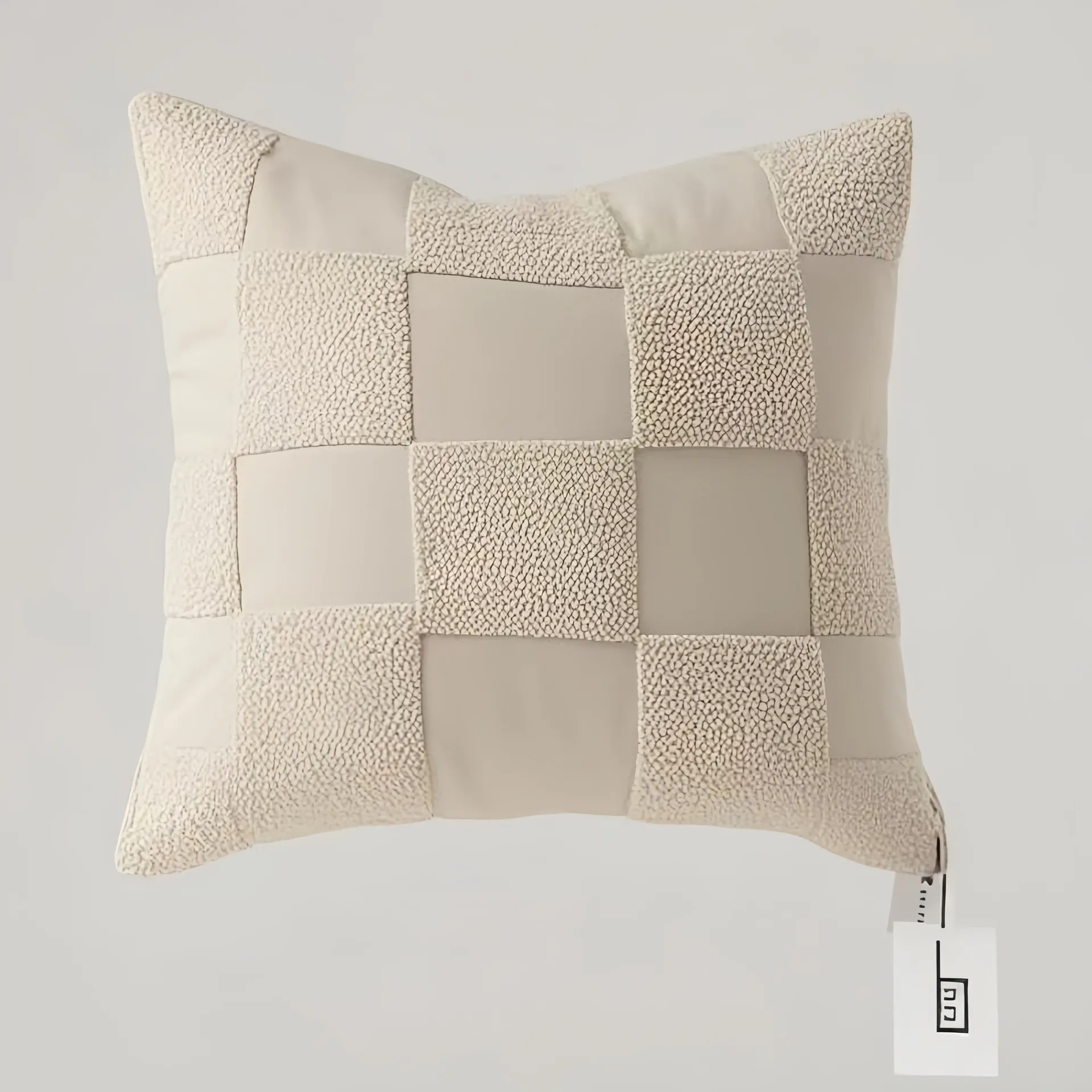A square beige pillow with a patchwork pattern made of textured and smooth fabric squares.
