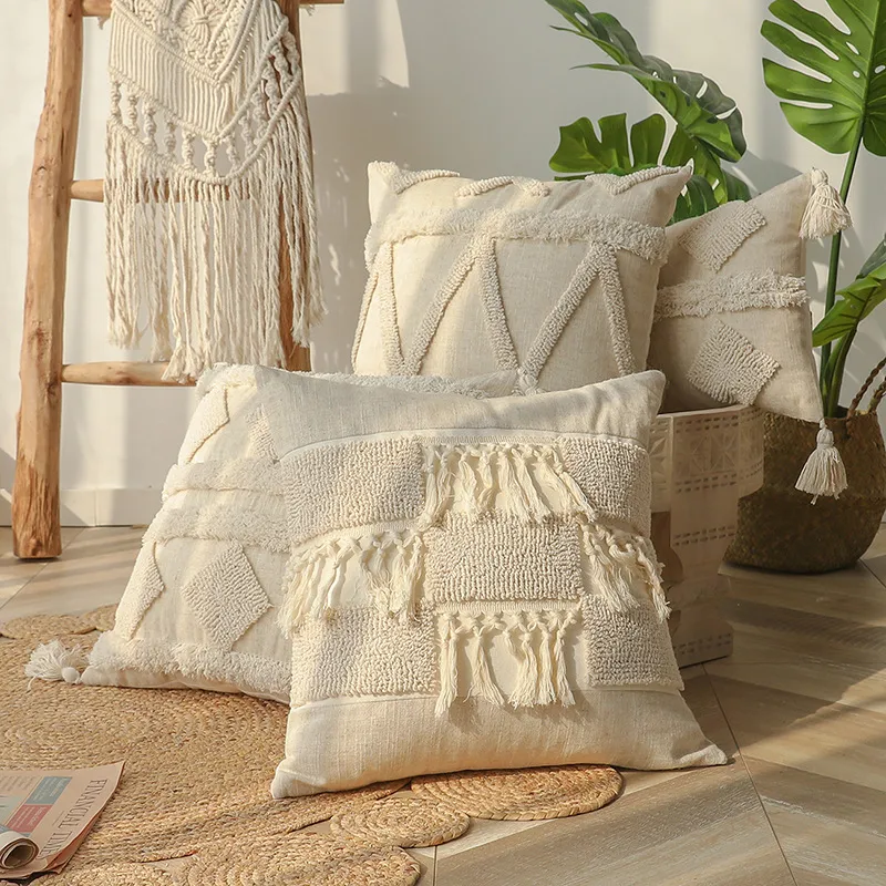 Trois coussins texturés aux motifs variés et ornés de pompons sont posés sur un tapis tissé. Derrière eux, on aperçoit une échelle en bois avec une suspension murale en macramé et une plante en pot.