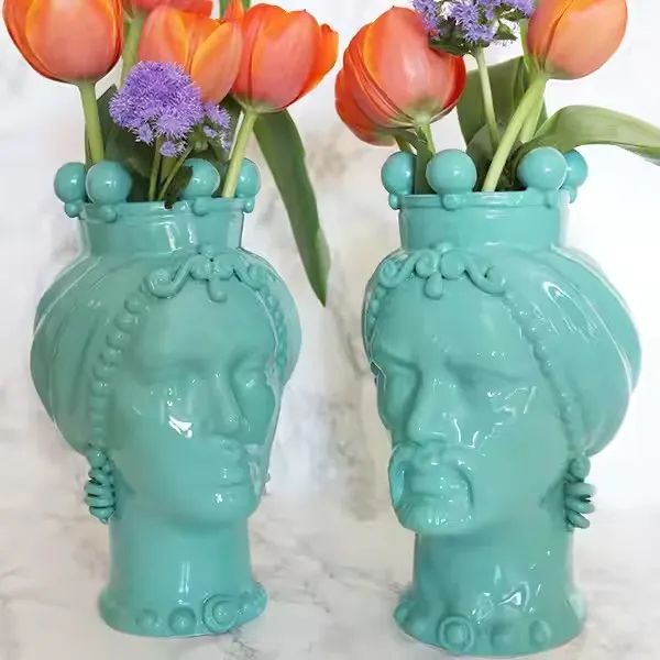 Two teal ceramic vases shaped like human faces filled with orange tulips and purple flowers.
