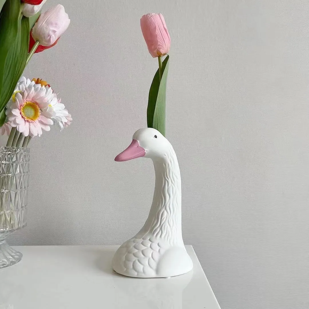 White ceramic swan vase holding a pink tulip, placed on a white surface next to a glass vase with mixed flowers.