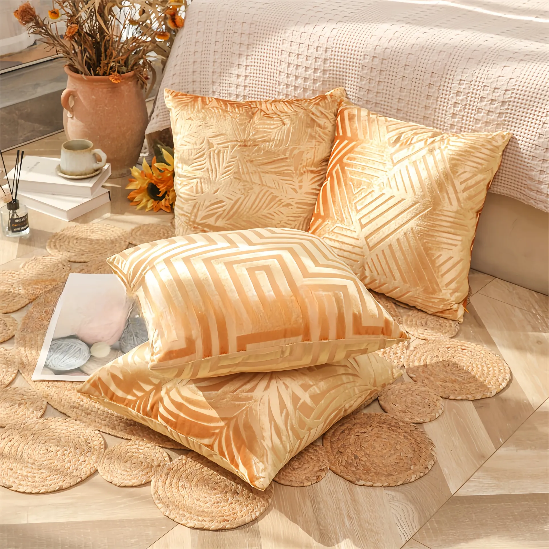Quatre coussins décoratifs à motifs géométriques posés sur un tapis tissé à côté d'un lit, entourés d'un vase, de livres et d'une tasse.