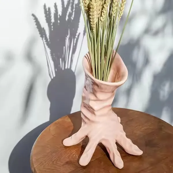 A unique ceramic vase shaped like a hand with fingers spread out, holding dried wheat stalks, casting an artistic shadow.