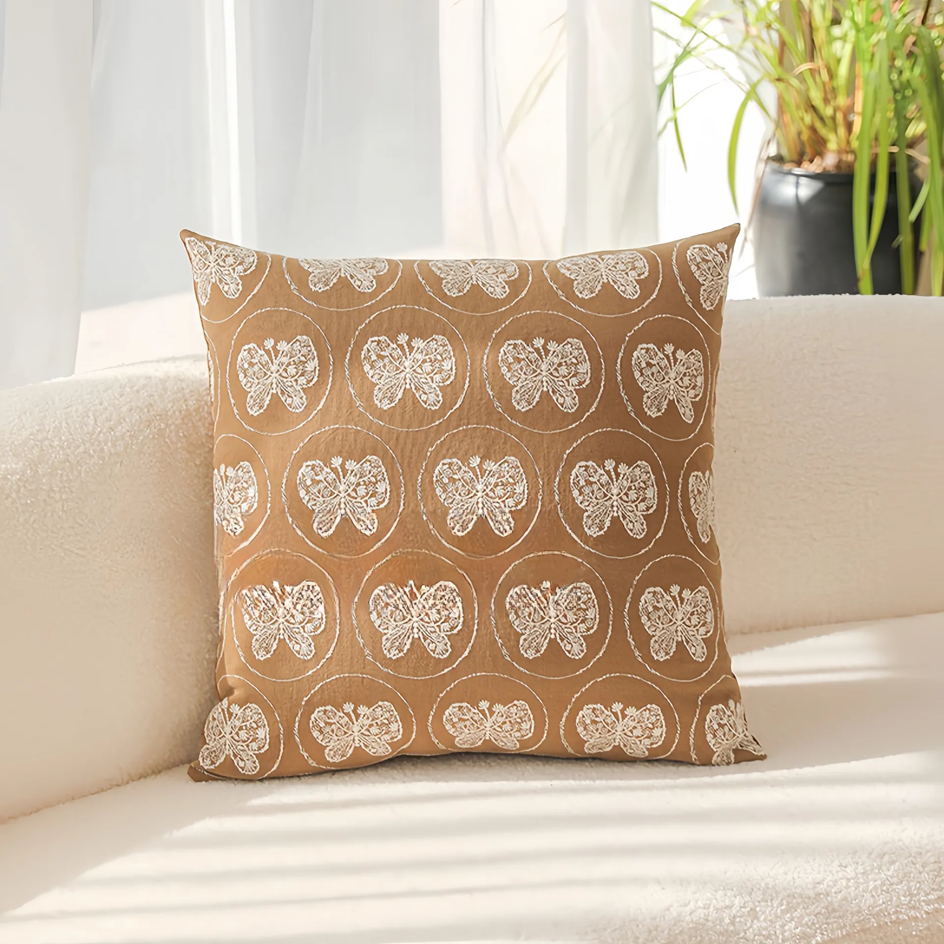 Un coussin décoratif marron avec des motifs de papillons blancs, placé sur un canapé couleur crème avec la lumière du soleil qui passe par une fenêtre.