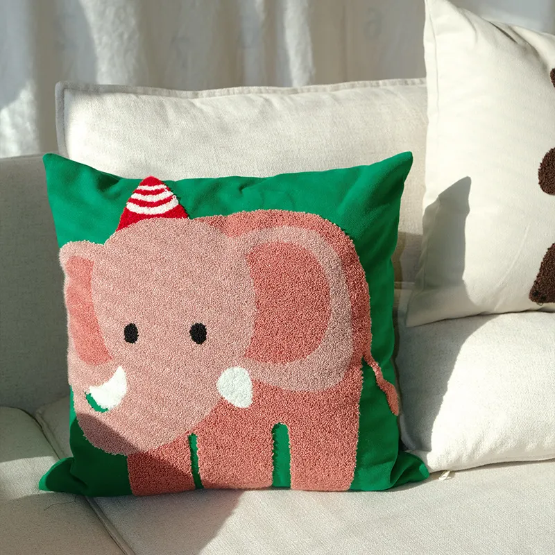 Un coussin décoratif avec un éléphant rose et un chapeau de fête rayé rouge et blanc, placé sur un canapé de couleur claire.