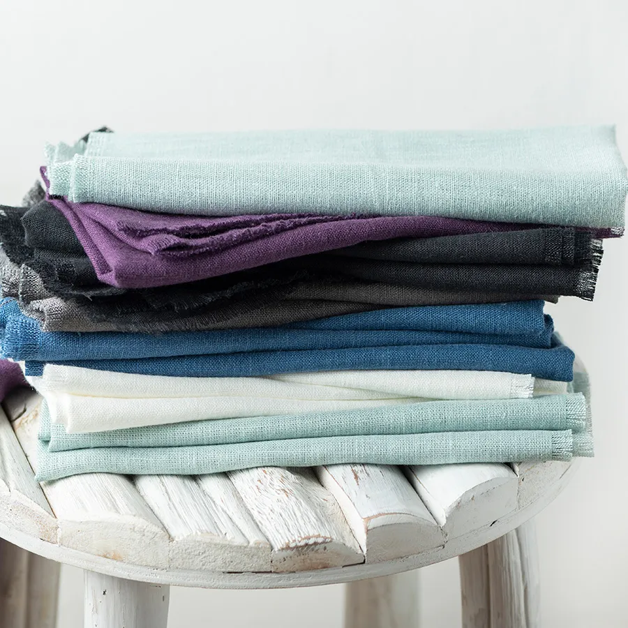 A stack of folded fabrics in various colors including teal, purple, grey, and blue, placed on a rustic white wooden stool.