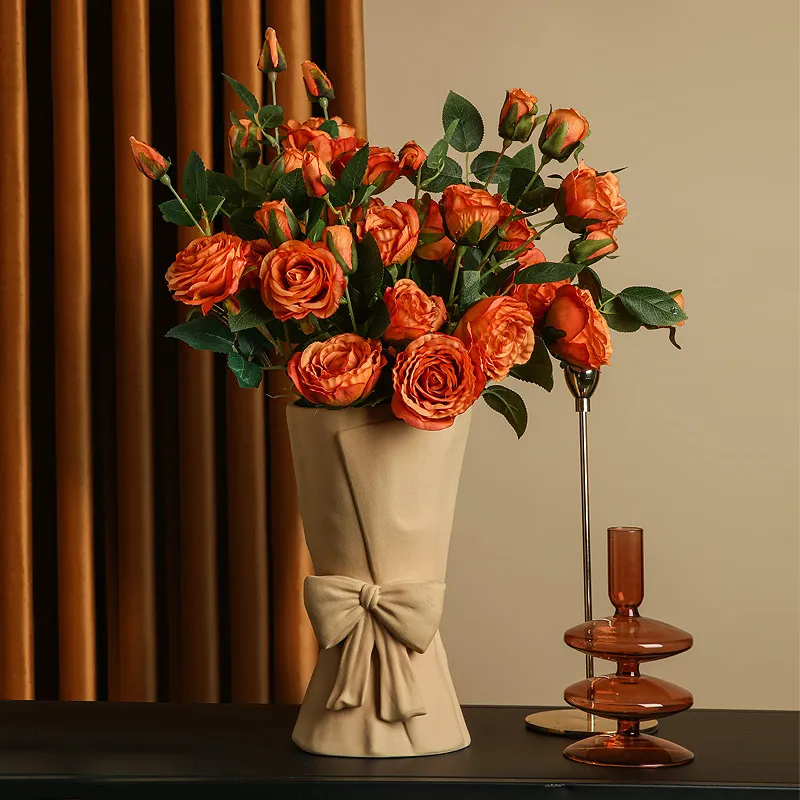 A bouquet of orange roses in a beige vase with a bow, placed on a table next to a brown decorative glass object.