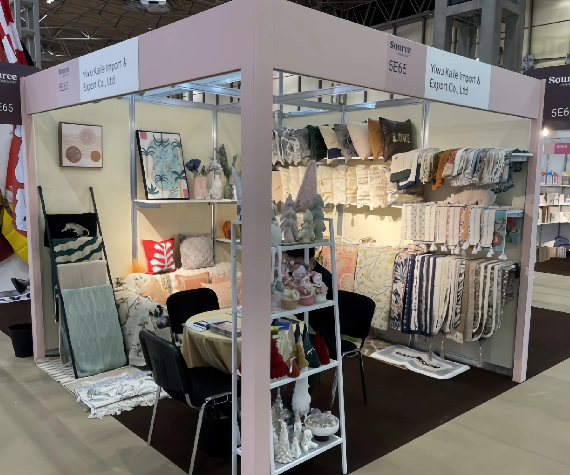 Trade show booth displaying decorative textiles and home items by Yiwu Kalee Import & Export Co., Ltd. The setup features shelves with various cushions, cloths, and small decorative items.