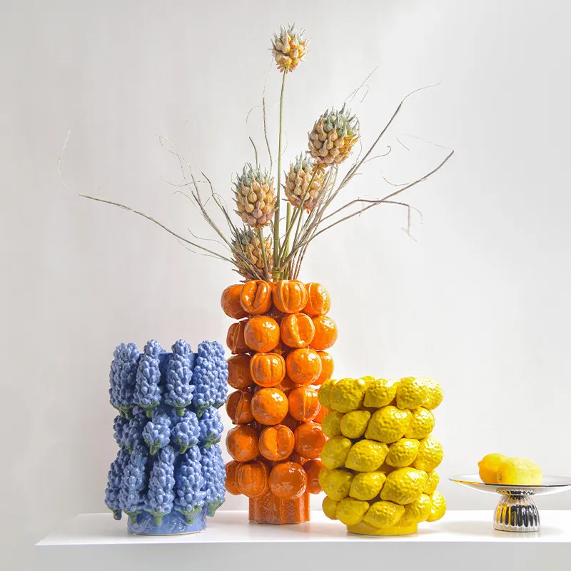 Three vases shaped like clusters of fruits in blue, orange, and yellow, with plants and a small plate of lemons on a white surface.