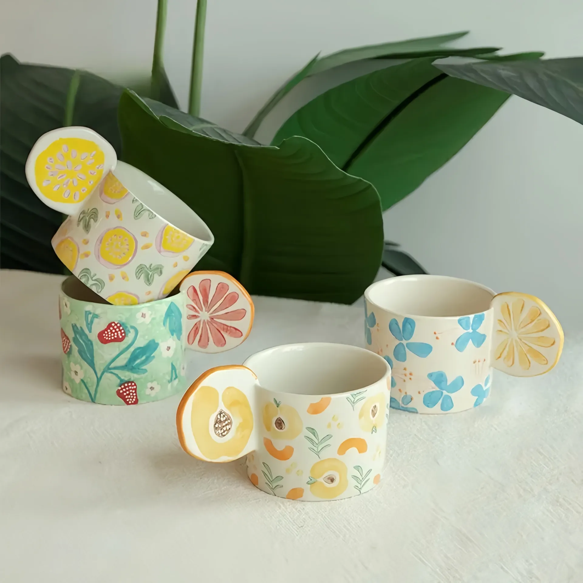 Four ceramic mugs with fruit-themed handles and colorful floral patterns, set against a backdrop of large green leaves.