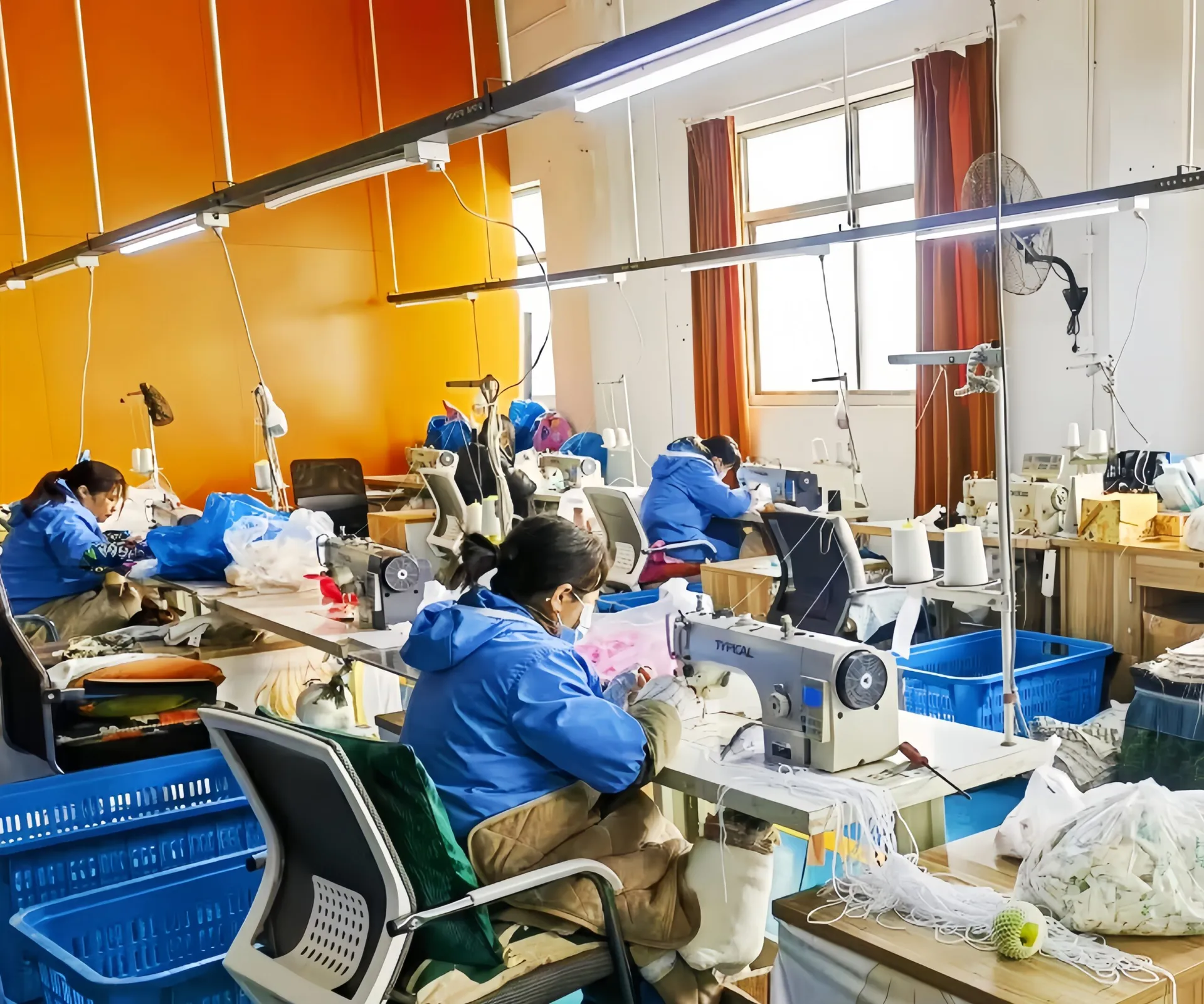 Workers in blue jackets sewing at machines in a garment factory with orange walls.