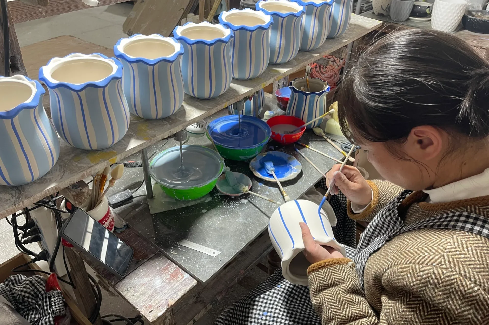 Pottery Painting Process A person painting blue stripes on pottery vases in a workshop filled with various paints and tools.